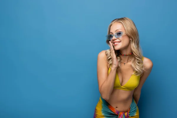 Усміхнена молода жінка в сонцезахисних окулярах дивиться на камеру ізольовано на синьому — стокове фото