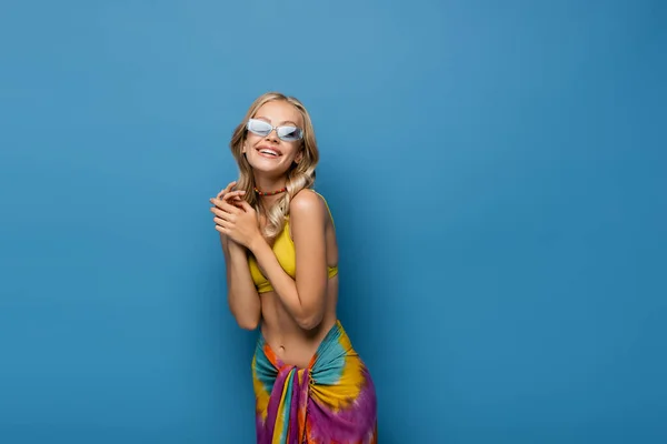 Радостная молодая женщина в солнечных очках и бисером красочное ожерелье улыбаясь изолированы на синий — стоковое фото