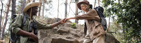 Smiling african american man holding hand of wife with backpack in forest, banner — Stock Photo