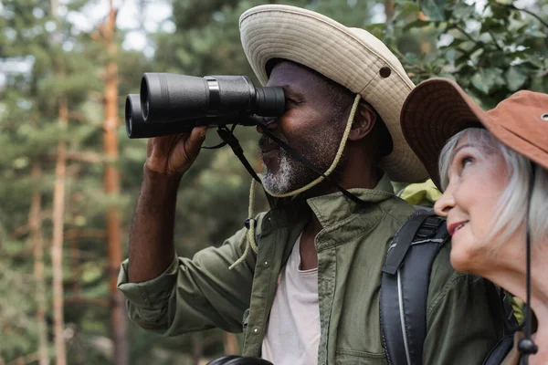 African american hiker looking though binoculars near smiling wife — Stock Photo