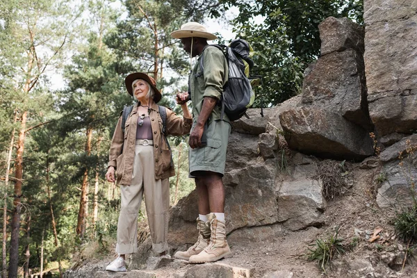 Elderly woman holding hand of african american husband near stones in forest — Stock Photo