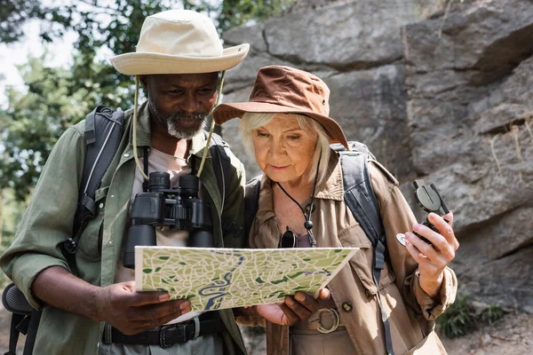 Pareja multiétnica de excursionistas con brújula mirando el mapa en el bosque - foto de stock