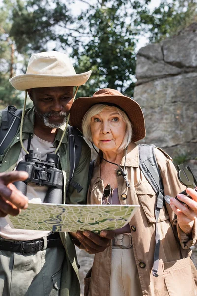 Senderistas multiétnicos mayores con brújula y mapa de pie en el bosque - foto de stock