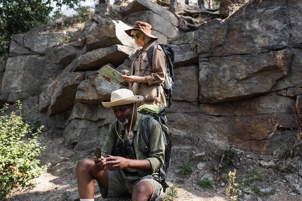 Senior multiethnic hikers using map and compass near rock in forest — Stock Photo