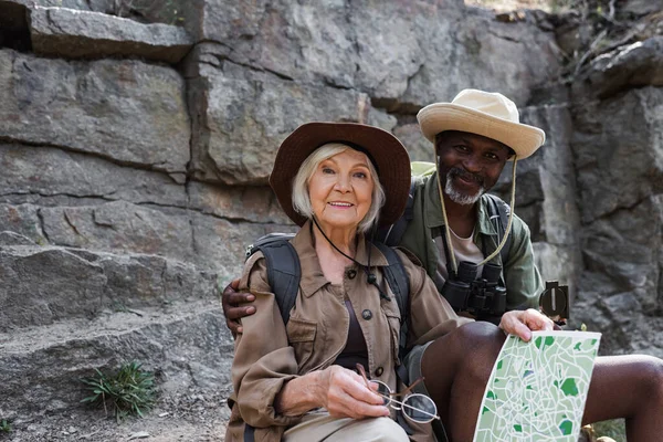 Afrikanischer Wanderer umarmt lächelnde Frau mit Rucksack und Karte in der Nähe von Felsen — Stockfoto