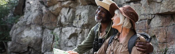 Senior afroamericano excursionista abrazando sonriente esposa con mapa cerca de roca, pancarta - foto de stock