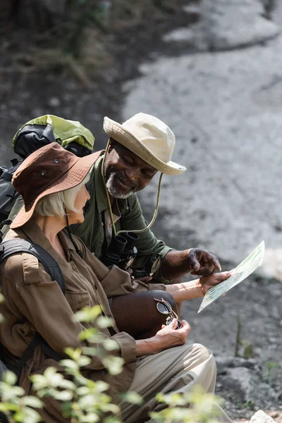 Caminhante afro-americano apontando para mapa perto da esposa sênior com óculos de sol na floresta — Fotografia de Stock