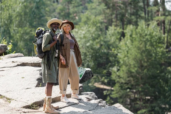 Cheerful multiethnic senior travelers with backpacks standing on rock — Stock Photo