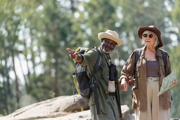 Smiling african american hiker pointing with finger near wife with map in forest — Stock Photo