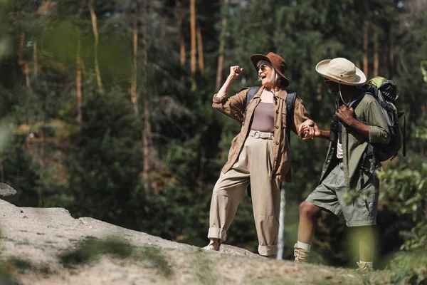 Excited elderly hiker showing yes gesture while holding hand of african american husband on rock — Stock Photo