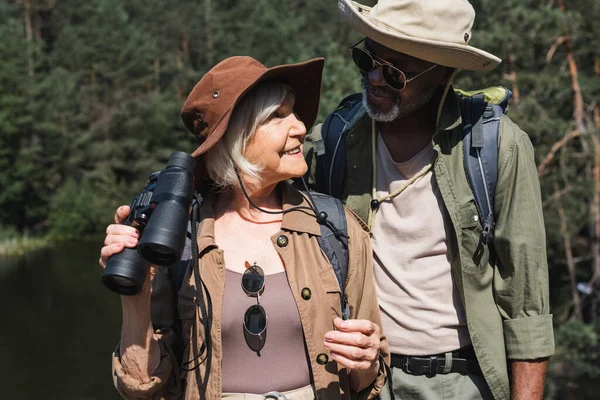 Viajante sênior feliz segurando binóculos perto do marido afro-americano na floresta — Fotografia de Stock