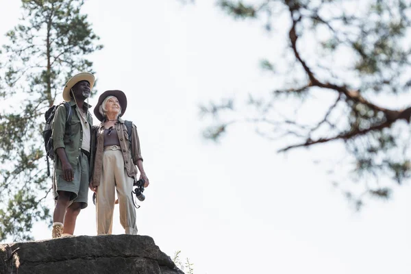 Low angle view of smiling interracial hikers with binoculars standing on rock — Stock Photo