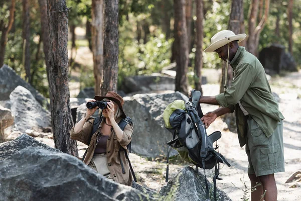 Africano americano homem segurando mochila perto da esposa com binóculos e pedras na floresta — Fotografia de Stock