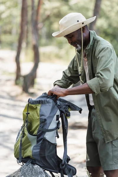 Elderly african american hiker in hat holding backpack near stone in forest — Stock Photo