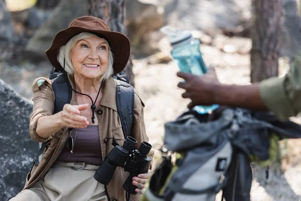 Elderly tourist with binoculars outreaching hand near african american husband with bottle of water in forest — Stock Photo