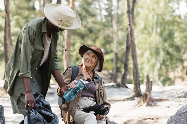 Африканський турист тримає пляшку води біля усміхненої дружини з біноклем у лісі. — стокове фото