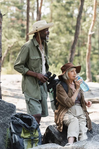 Sonriente excursionista afroamericano sosteniendo prismáticos cerca de esposa mayor con botella de agua en piedras - foto de stock