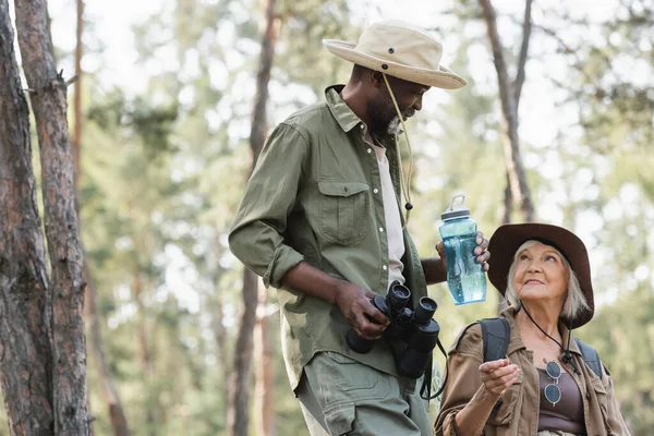 Senior woman talking to african american husband with water and binoculars in forest — Stock Photo