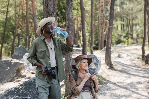 African american traveler holding bottle of water and binoculars near smiling wife in forest — Stock Photo