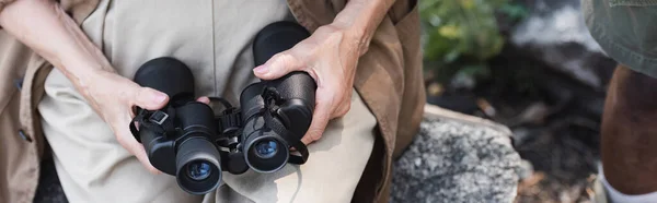 Cropped view of elderly hiker holding binoculars on stone in forest, banner — Stock Photo