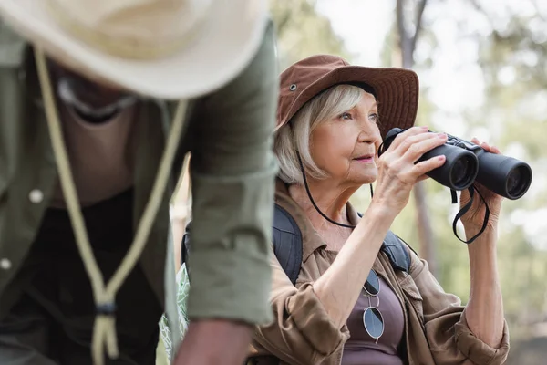 Senior tourist with binoculars looking away near african american husband on blurred foreground — Stock Photo