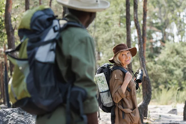 Lächelnde ältere Frau mit Rucksack und Fernglas schaut verschwommenen afrikanisch-amerikanischen Ehemann im Wald an — Stockfoto