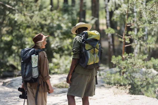 Positive hiker with binoculars looking at african american husband in forest — Stock Photo