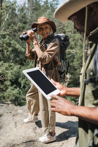 Lächelnde Frau mit Fernglas neben verschwommenem afrikanisch-amerikanischem Ehemann mit digitalem Tablet im Wald — Stockfoto