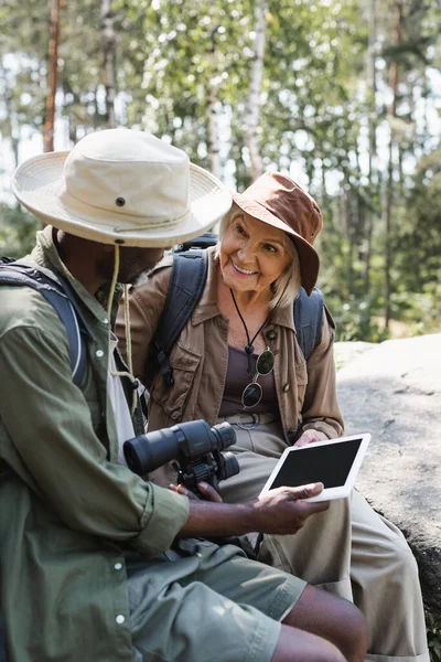 Positive traveler looking at african american husband with binoculars and digital tablet in forest — Stock Photo