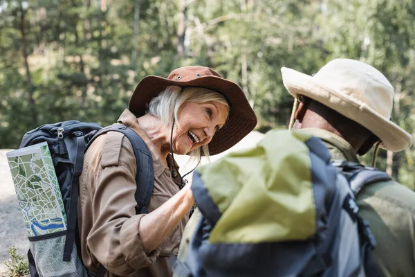 Happy senior hiker with backpack and mal looking at blurred african american husband in forest — Stock Photo