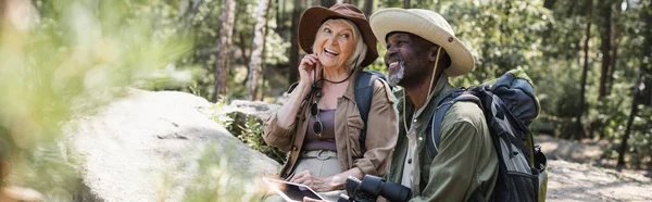 Cheerful multiethnic hikers with binoculars and digital tablet in forest, banner — Stock Photo