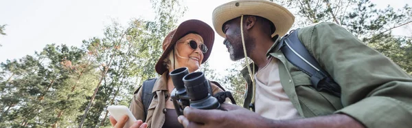 Low angle view of positive interracial tourists with smartphone and binoculars in forest, banner — Stock Photo
