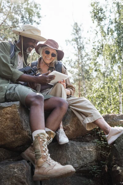 Randonneurs multiethniques souriants avec jumelles et tablette numérique assis sur des pierres dans la forêt — Photo de stock