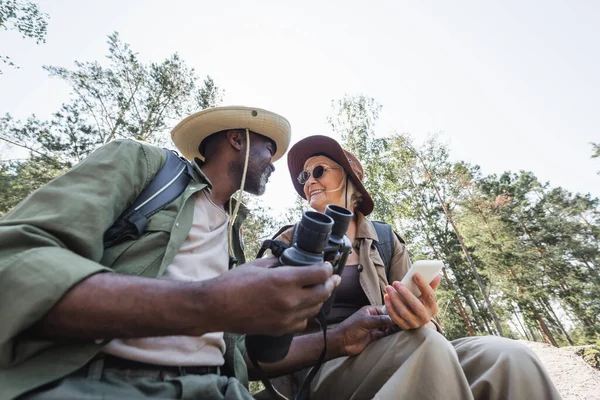 Tiefansicht positiver multiethnischer Wanderer mit Smartphone und Fernglas im Wald — Stockfoto