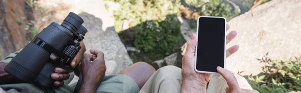 Top view of interracial couple holding smartphone and binoculars on stones, banner — Stock Photo