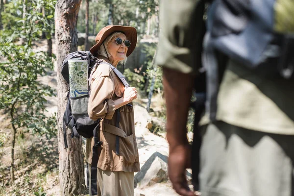 Mujer positiva con mochila y mapa de pie en el bosque cerca borrosa marido afroamericano - foto de stock