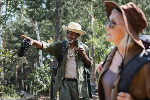 Smiling african american man with binoculars pointing with finger near blurred wife in forest — Stock Photo