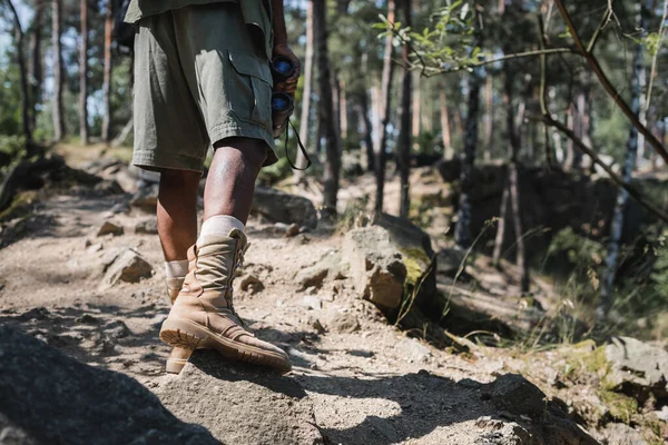 Cropped view of african american traveler holding binoculars on path with stones in forest — Stock Photo