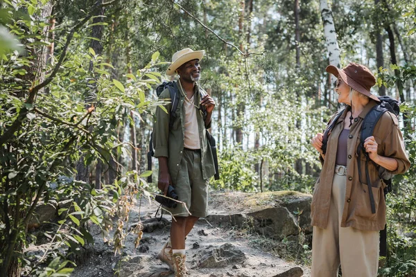 Casal idoso interracial com mochilas sorrindo enquanto caminhava na floresta — Fotografia de Stock