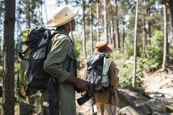 Afrikanischer Wanderer mit Rucksack und Fernglas wandert nahe verschwommener Frau im Wald — Stockfoto