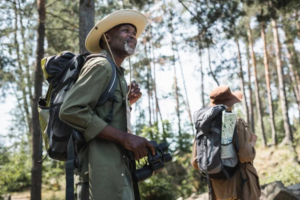 Улыбающийся африканский американец, держащий бинокль во время похода с женой в лесу — стоковое фото