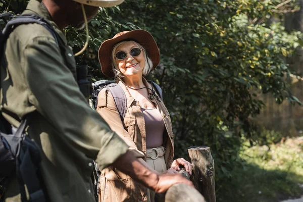 Lächelnde Frau mit Sonnenbrille blickt Afroamerikaner mit Rucksack in der Nähe von Zaun im Wald an — Stockfoto