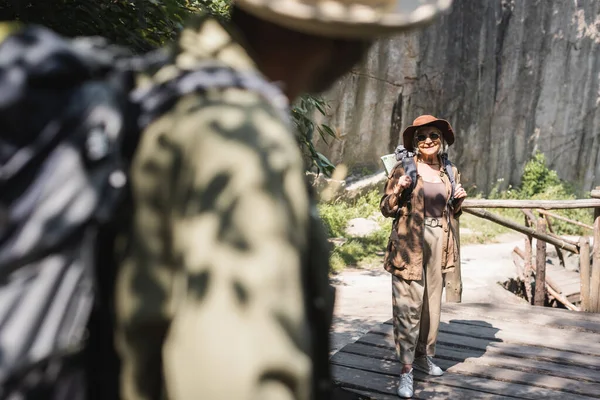 Senior traveler with backpack smiling at blurred african american husband on bridge — Stock Photo