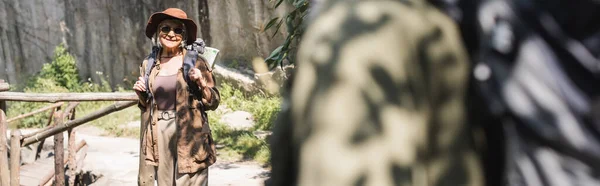 Mujer sonriente con mochila de pie cerca de marido borroso en el bosque, pancarta - foto de stock