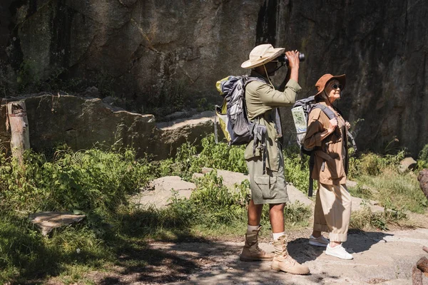 Homme afro-américain à la recherche de jumelles près de la femme avec sac à dos et falaise — Photo de stock