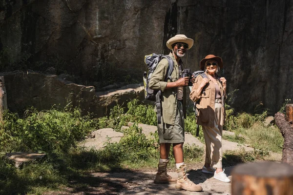 Sorrindo casal interracial com mochilas e binóculos olhando para a câmera perto do penhasco — Fotografia de Stock