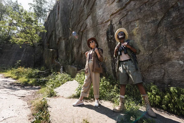 Interracial senior couple of travelers with backpacks and sunglasses standing near cliff — Stock Photo