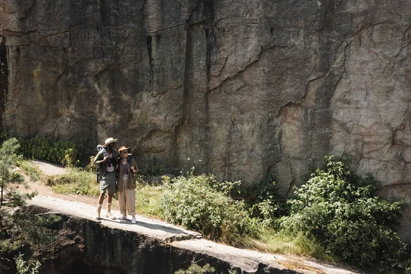 Межрасовая старшая пара с рюкзаками, идущая по тропинке рядом со скалой — стоковое фото