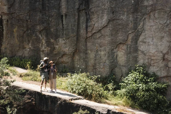 Pareja mayor interracial con mochilas caminando por el camino cerca del acantilado - foto de stock