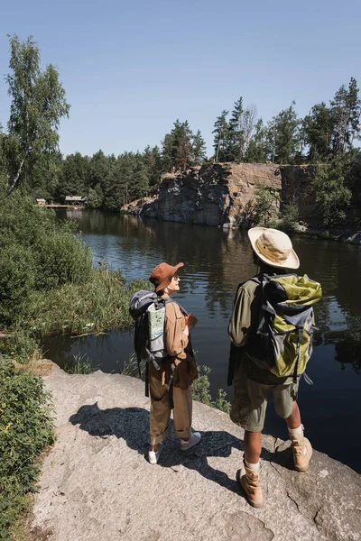 Smiling senior woman with backpack and map looking at african american husband near lake — Stock Photo